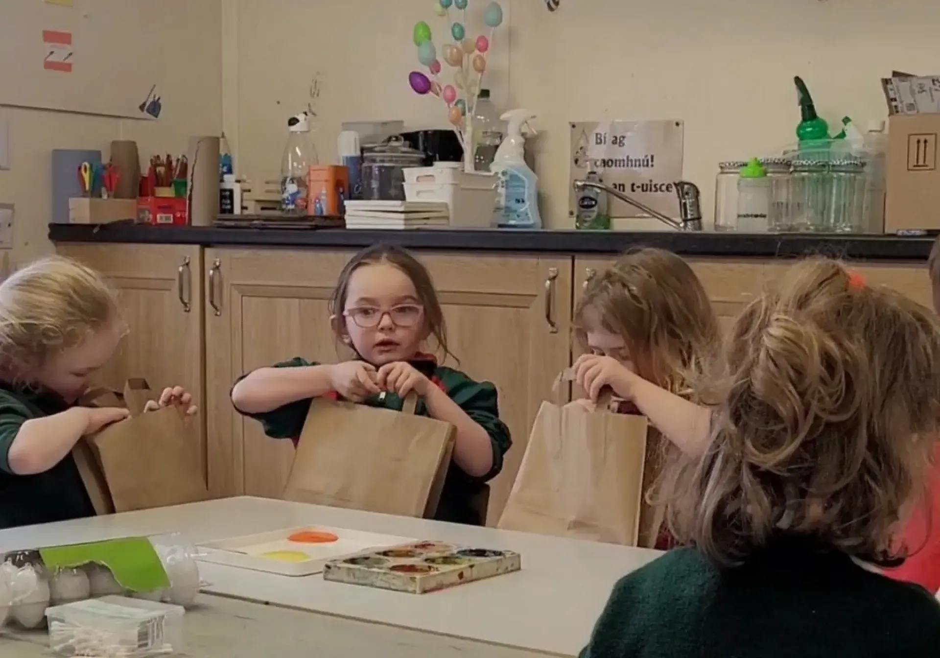 Young children seated at a table opening brown paper Bookbag packages.