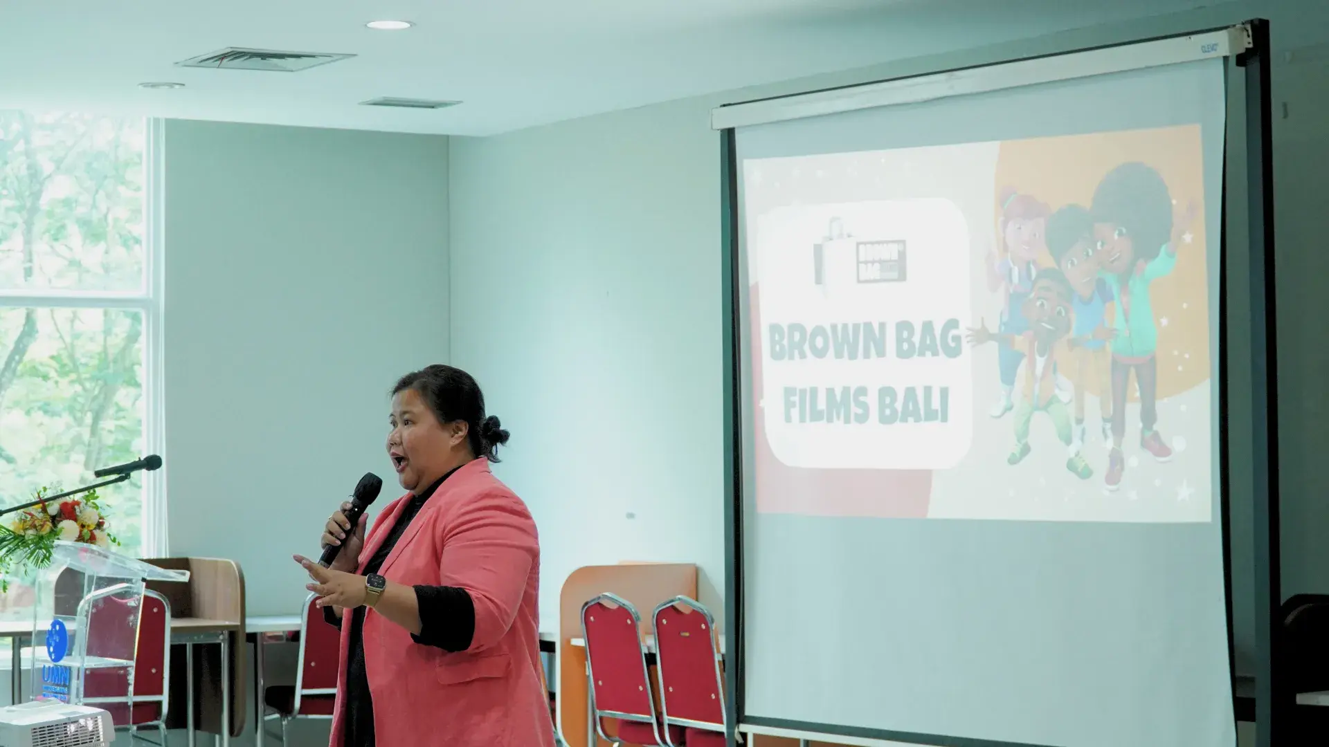 Person speaking into a microphone during a presentation with a screen showing Brown Bag Films Bali logo and animated characters.