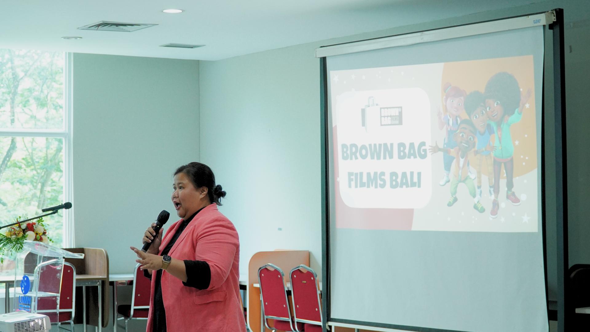 Person speaking into a microphone during a presentation with a screen showing Brown Bag Films Bali logo and animated characters.