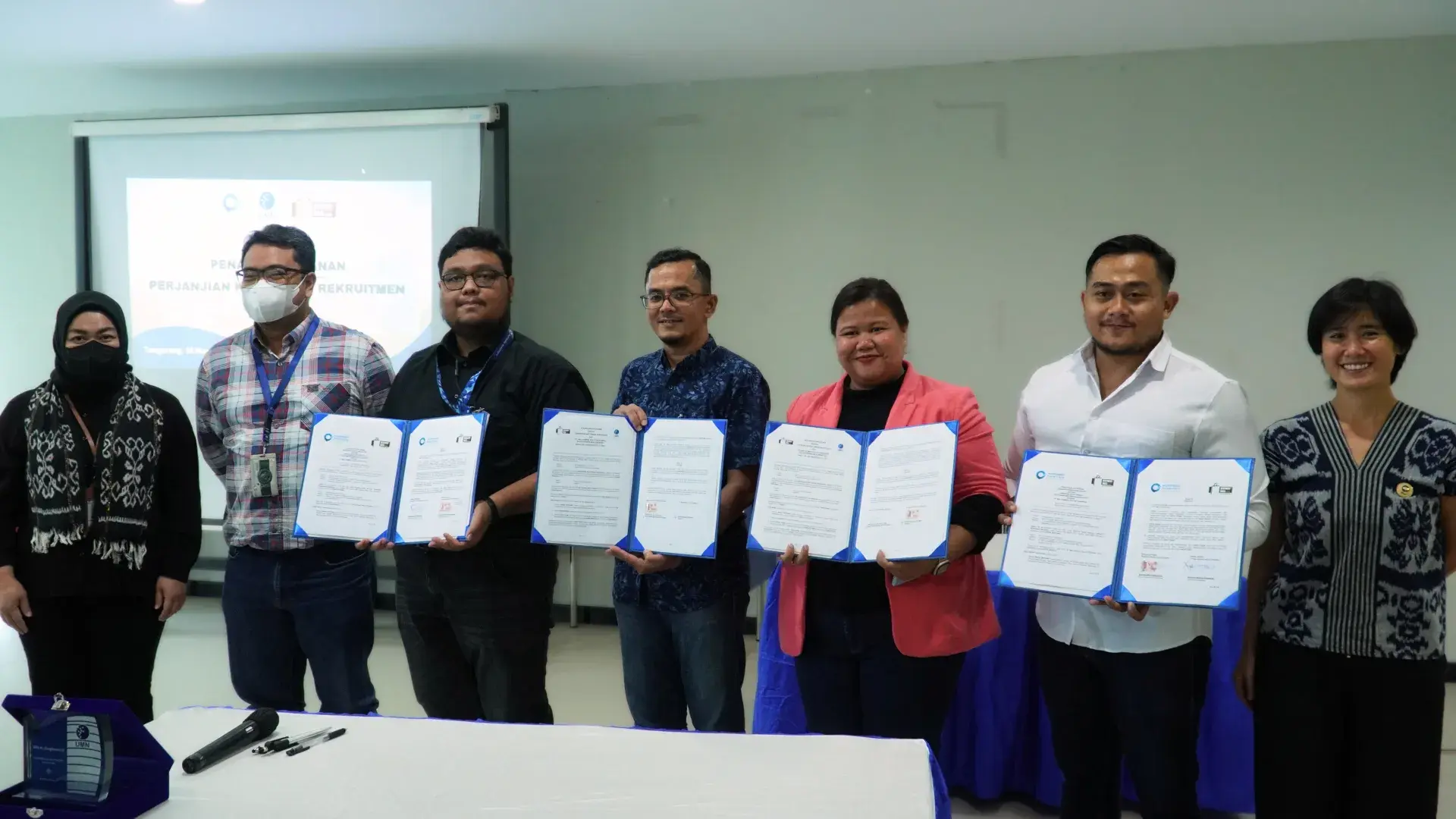 Group of people holding signed documents in front of a presentation screen during a formal agreement session.