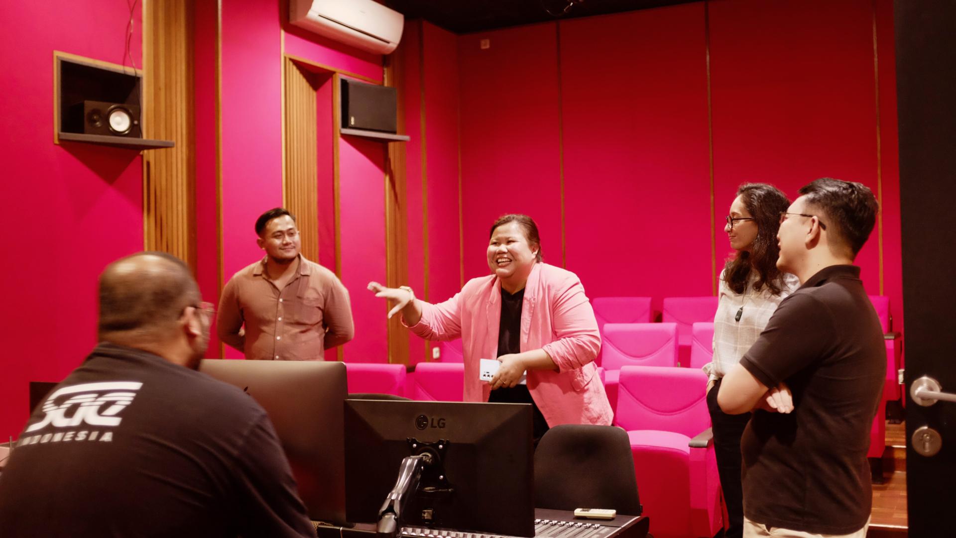 Group of people standing in a room with bright pink walls during a discussion near computer equipment.