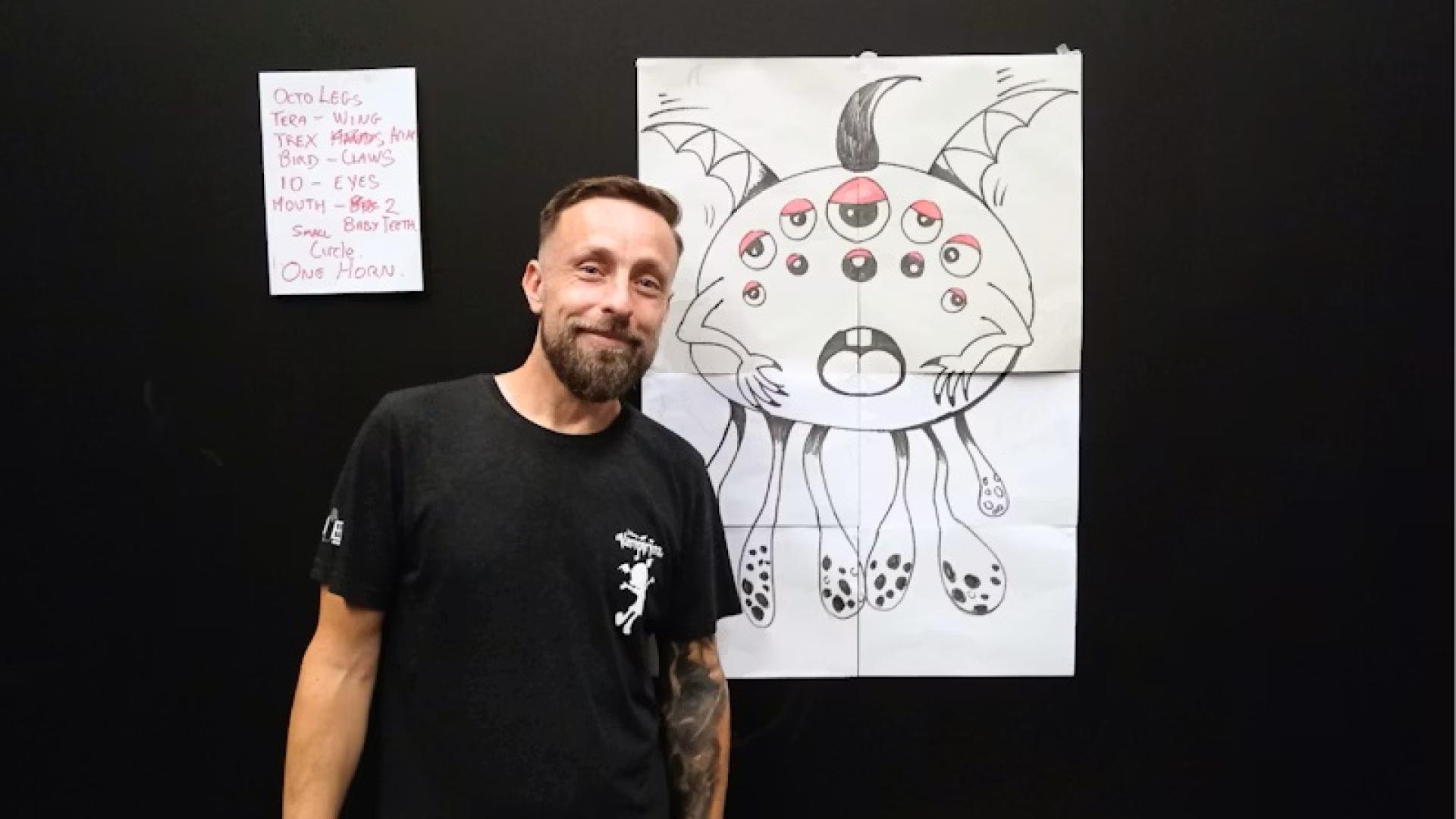 Person standing in front of a large black-and-white monster drawing with descriptive notes on the wall.