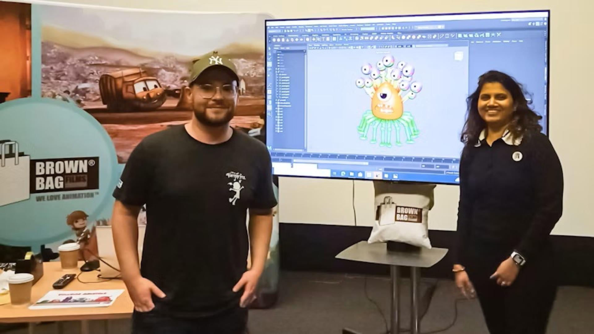Two people standing near a screen displaying a 3D monster model in an animation workshop.