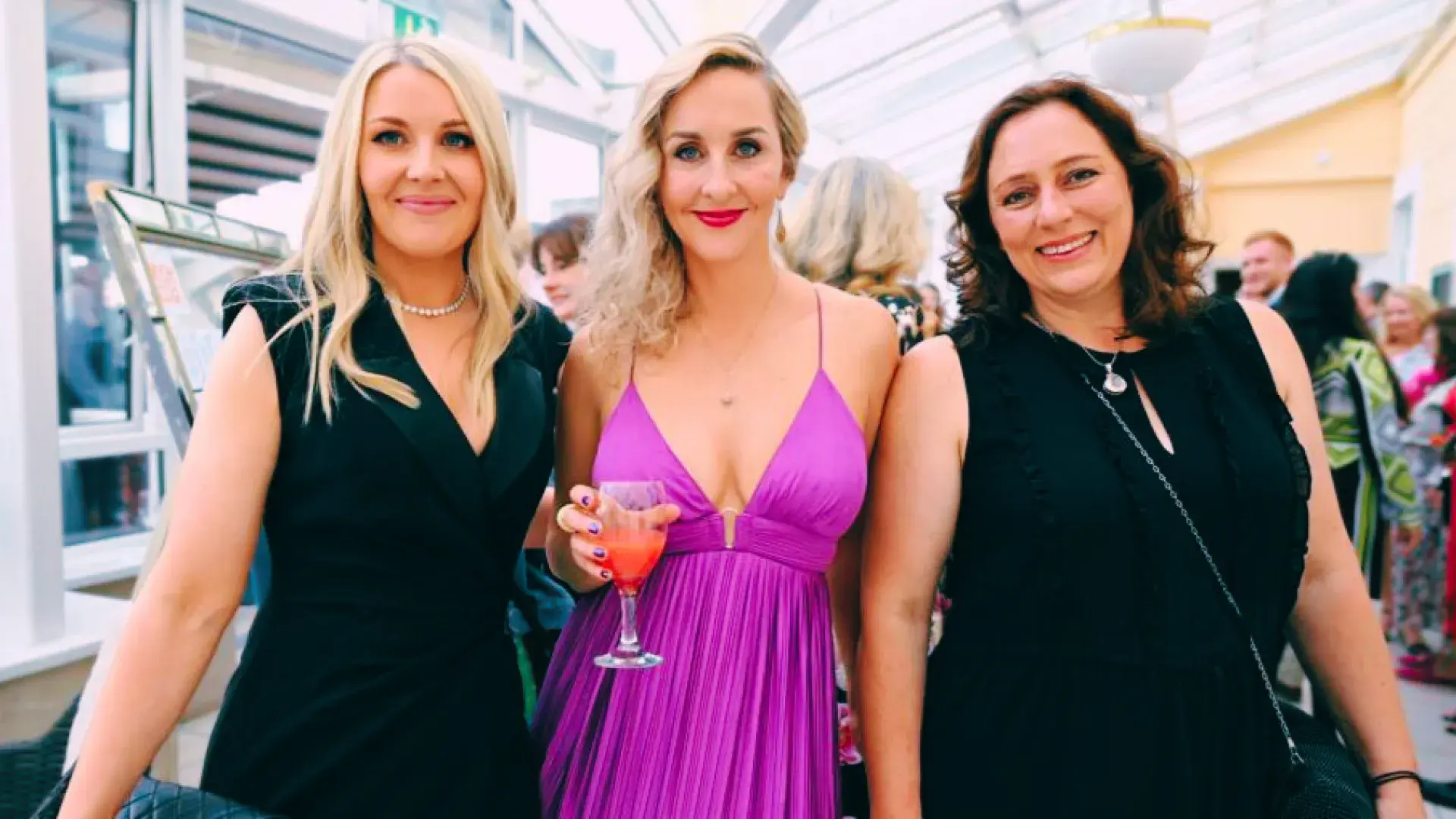 Three people standing indoors, one holding a drink, dressed in formal attire.