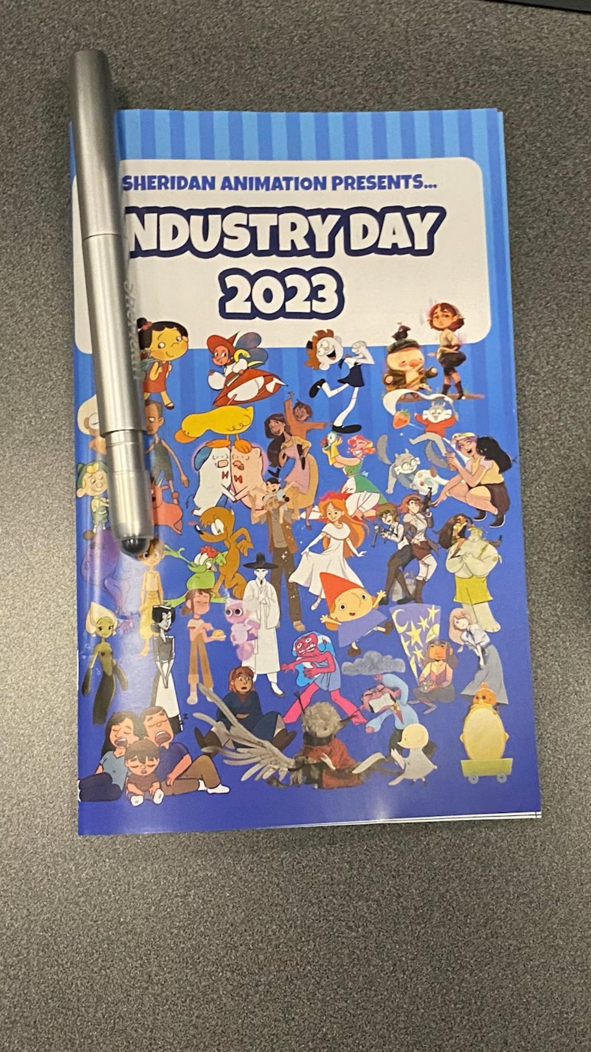 Industry Day 2023 booklet featuring cartoon characters on a blue cover with a silver pen placed on top.
