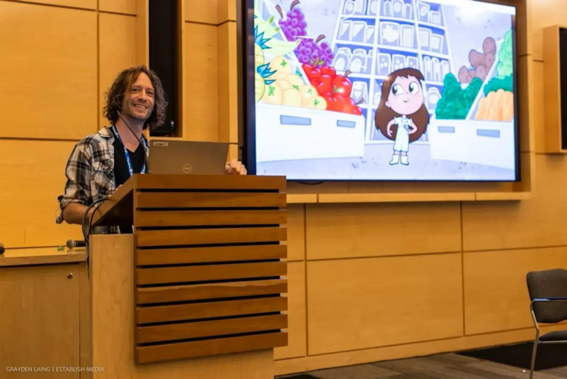 Speaker presenting at a podium with an animated character and grocery store scene displayed on a large screen.