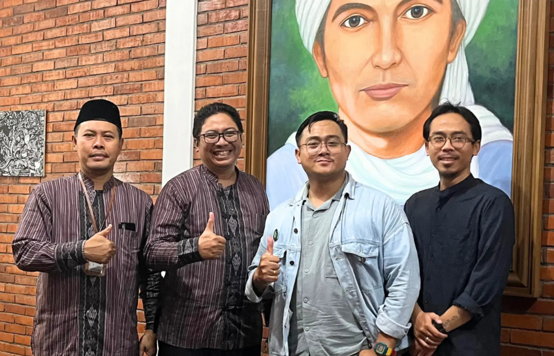 Four people standing in front of a brick wall and a large portrait painting, giving thumbs-up gestures.