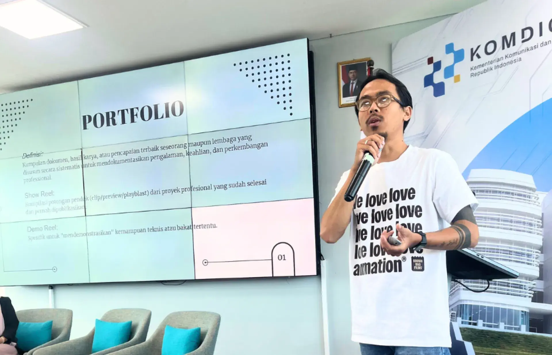 Presenter holding a microphone speaking in front of a screen displaying the word “PORTFOLIO” and presentation slides.