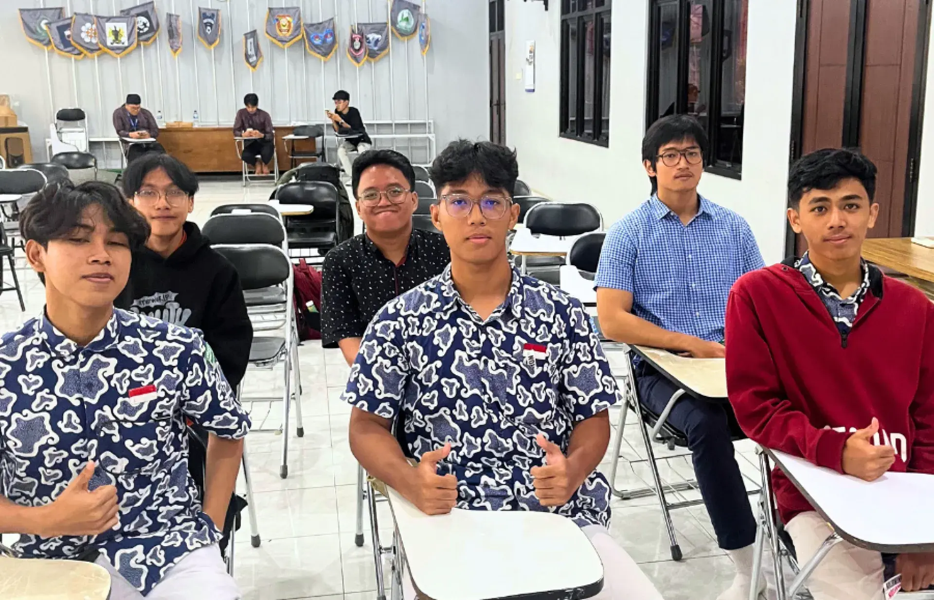 Students seated at desks in a classroom, several giving thumbs-up; banners displayed along the wall behind.