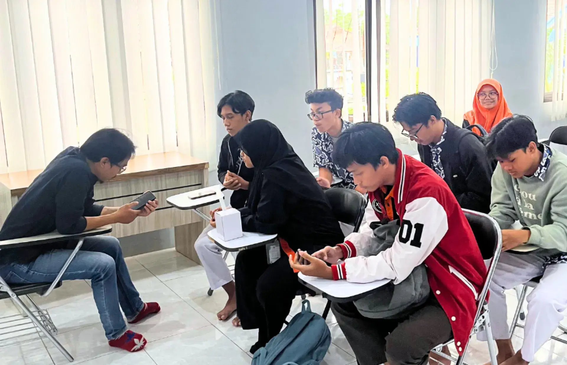 Small group of students seated in a classroom using phones and writing; windows with vertical blinds along the wall.