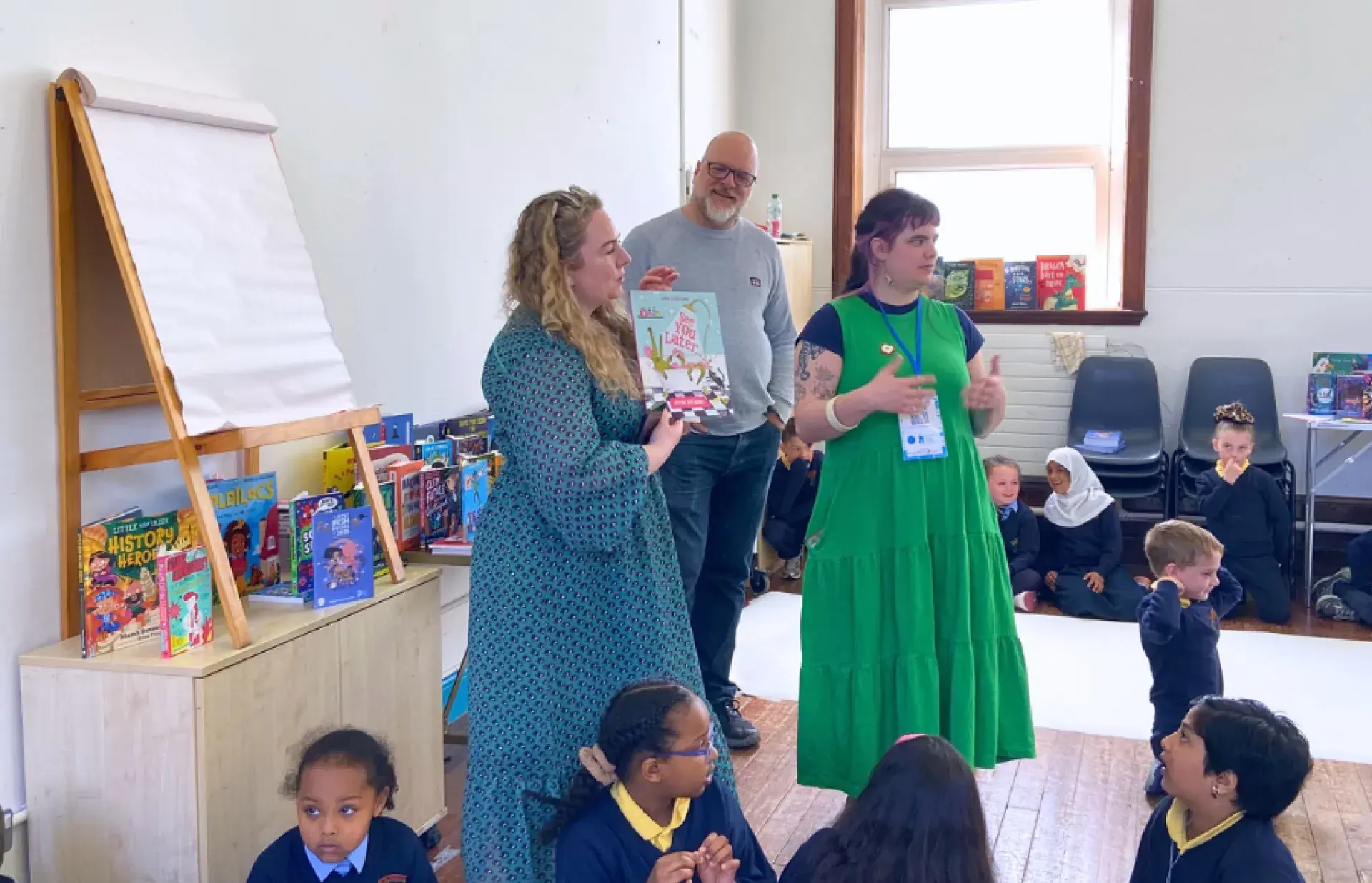 Adults presenting a children’s book to a seated group of schoolchildren in a classroom, with book displays along the walls.