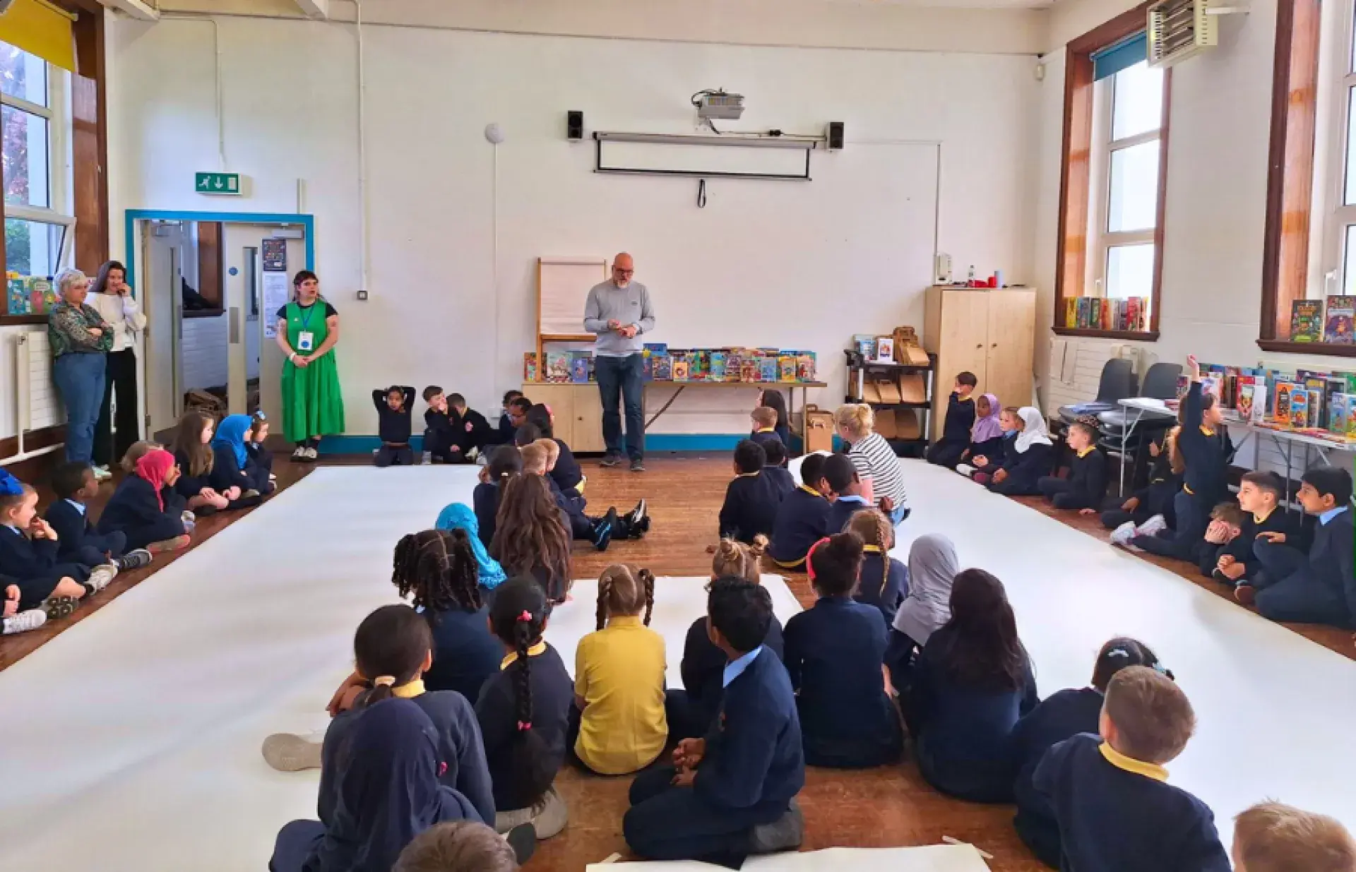 Wide view of a classroom where schoolchildren sit around on the floor while an adult stands at the front near children’s book displays.