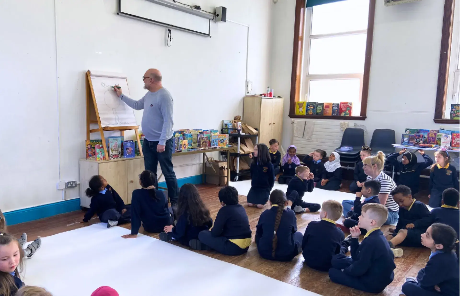 Person drawing on a flip chart at the front of a classroom while schoolchildren sit on the floor watching, with books displayed along the wall.
