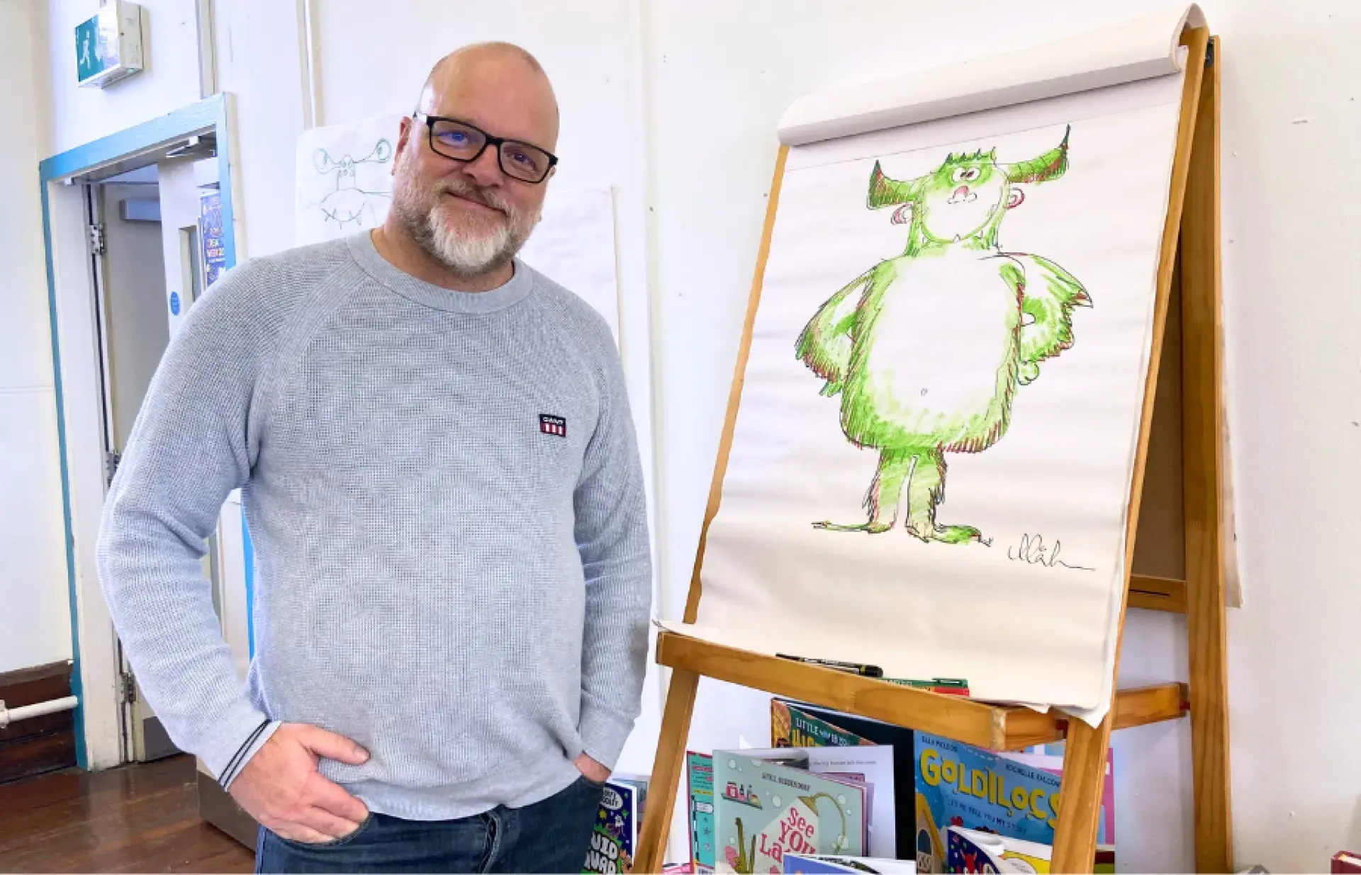 Person standing beside a flip chart showing a green monster illustration, with children’s books displayed on shelves below.