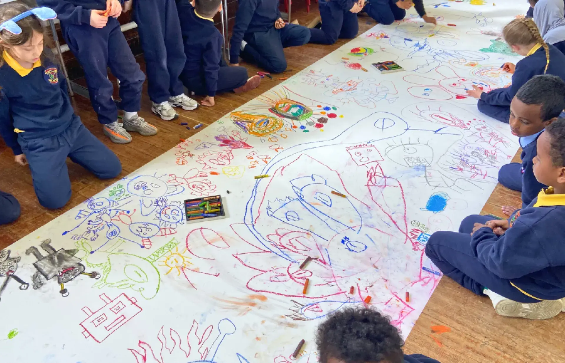 Schoolchildren seated and kneeling on the floor around a large sheet of paper covered in colourful drawings, with crayons and pencils scattered across the surface.