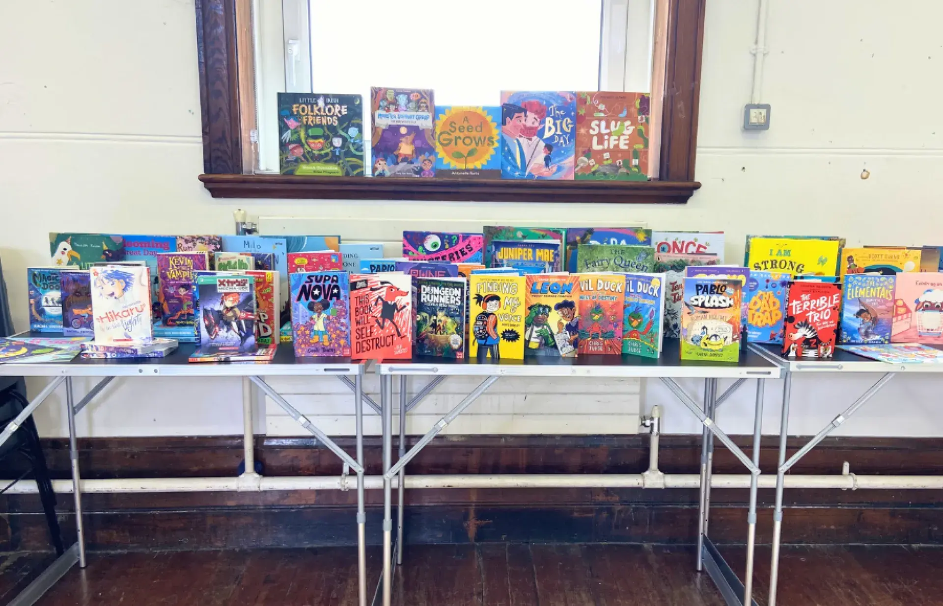 Children’s books displayed on fold‑out tables beneath a window, featuring brightly illustrated covers in a well‑lit room.