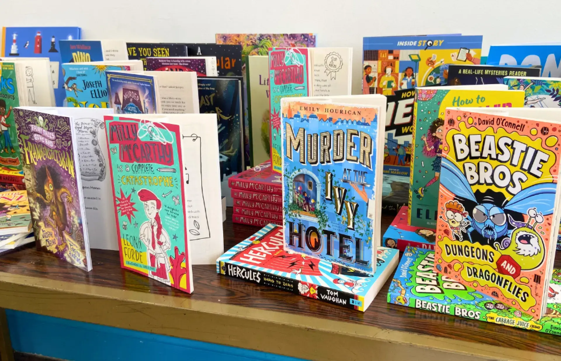 Colourful children’s books displayed upright on a wooden table, including mystery, comedy, and fantasy titles with illustrated covers.