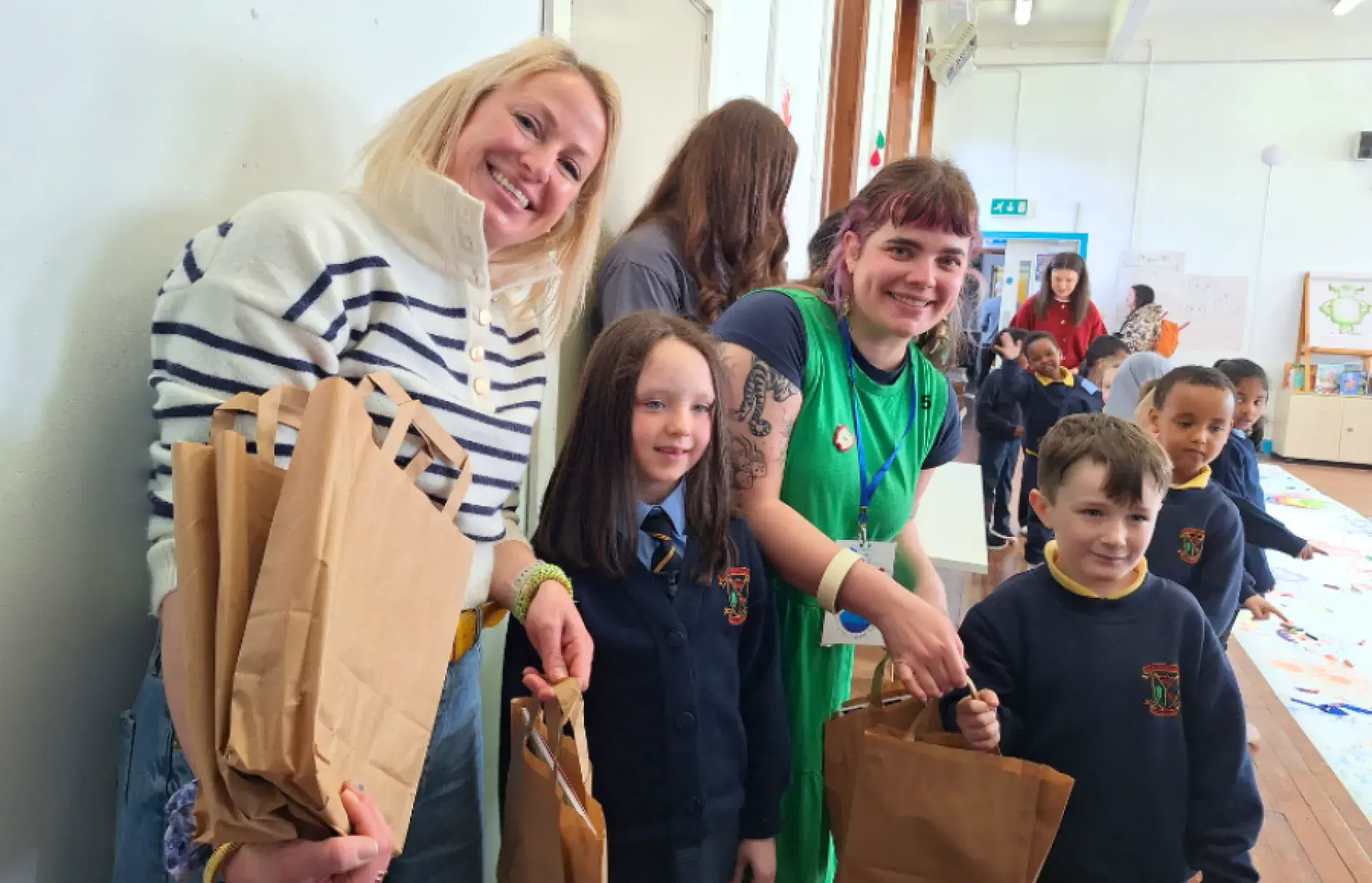 Adults and schoolchildren standing indoors, each holding brown paper bags, with more children and artwork visible in the background.