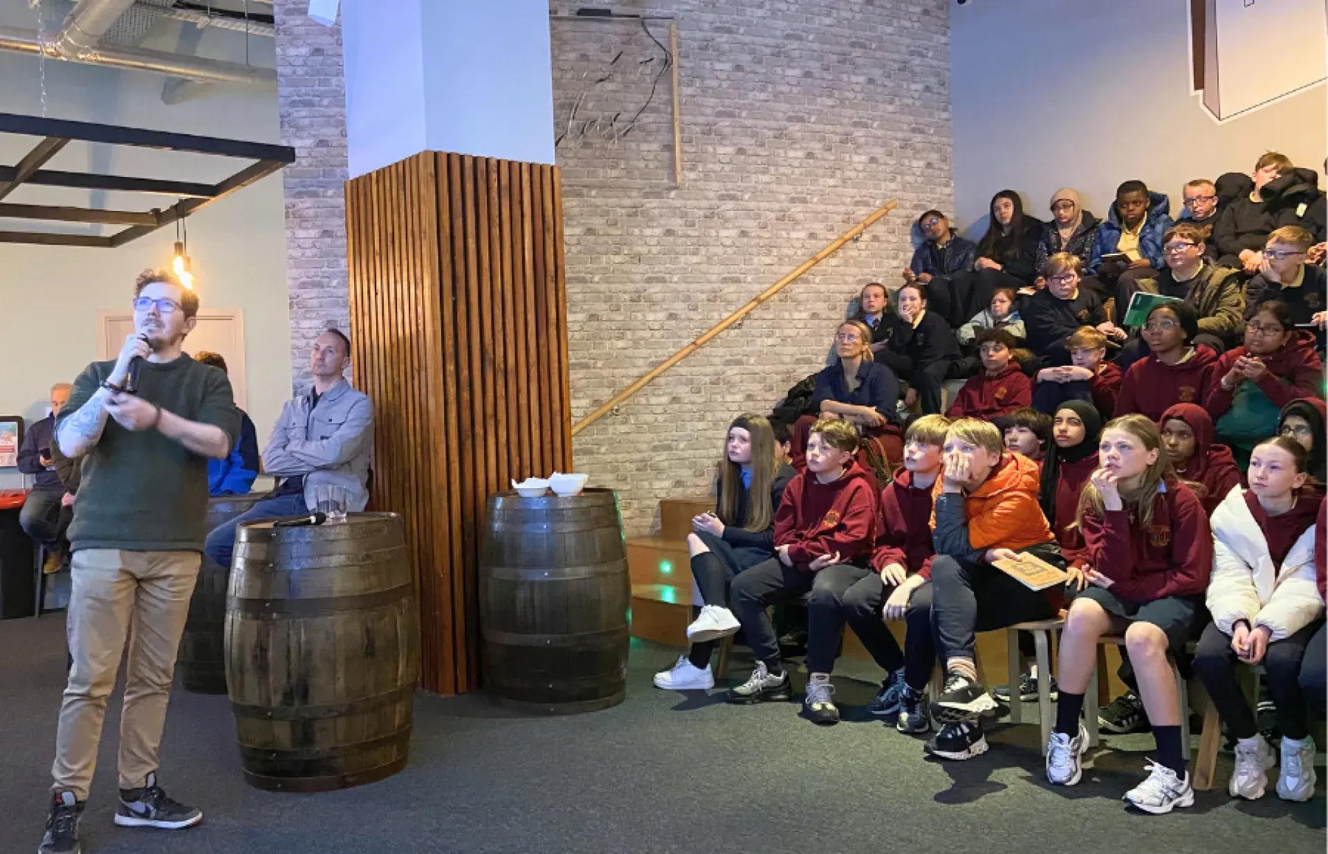 Adult holding a microphone and speaking to seated schoolchildren in a stepped seating area inside a brick‑walled room.