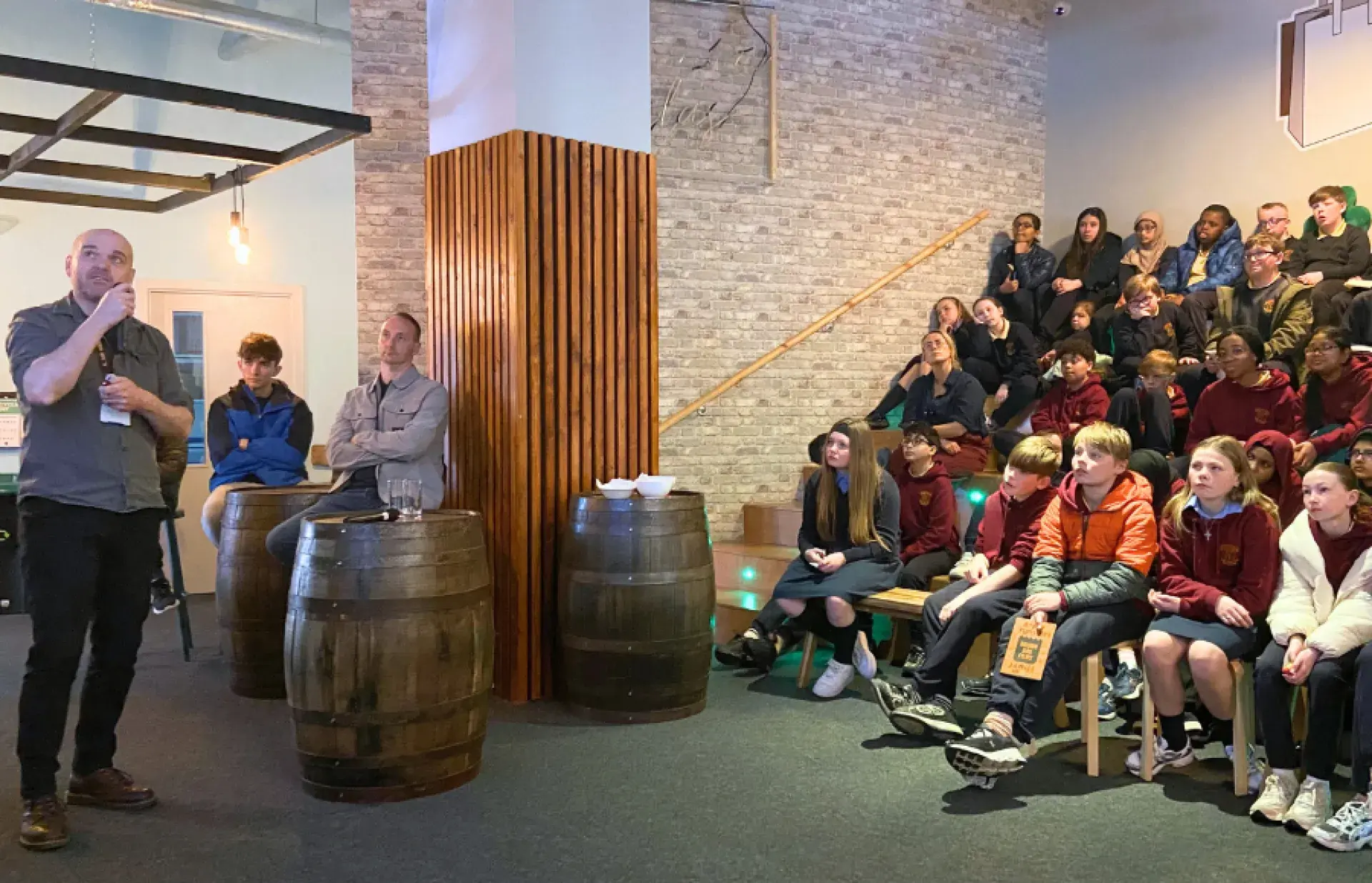 Adult speaking into a microphone beside seated schoolchildren arranged on stepped benches in an indoor venue.