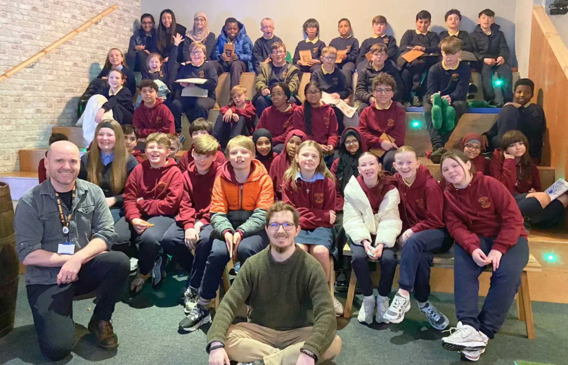 Large group of schoolchildren seated on tiered steps indoors, with two adults seated in front of the group.