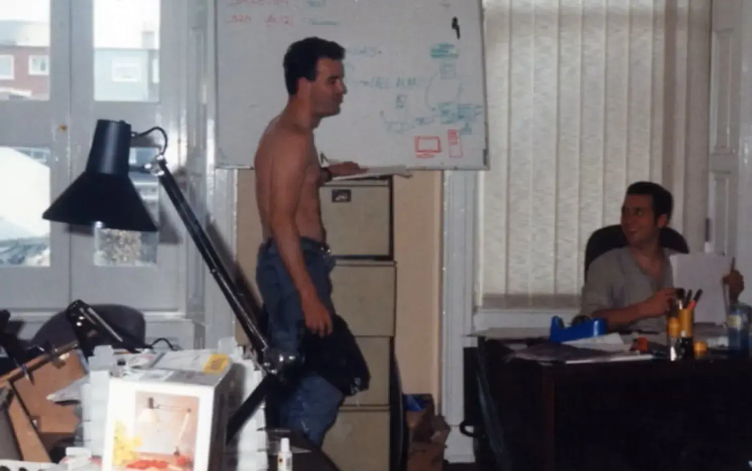 One person standing by a desk with computer equipment, another seated nearby, both working.