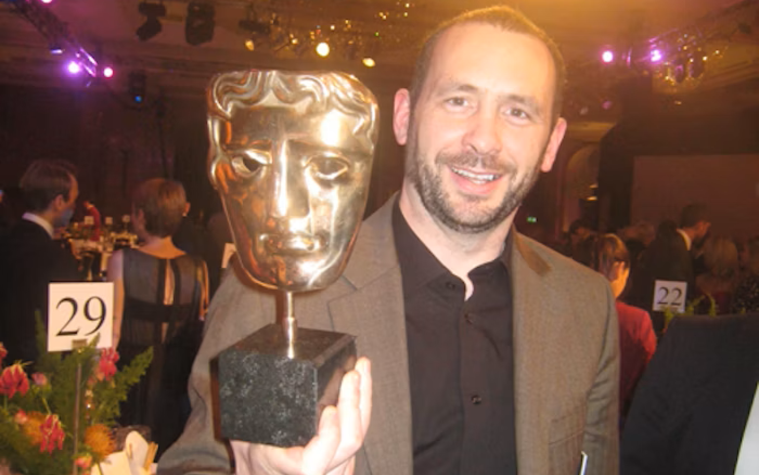 A person in a brown suit jacket smiling while holding a BAFTA award in a warmly lit banquet hall with tables, floral centerpieces, and chandeliers.