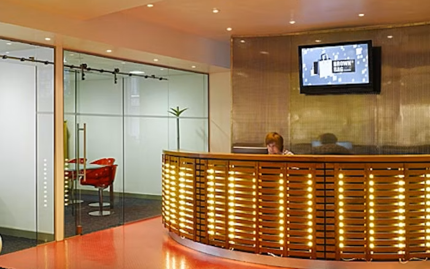 Modern office reception with a curved wooden desk, red tile flooring, recessed lighting, and a glass-walled meeting room with red chairs.