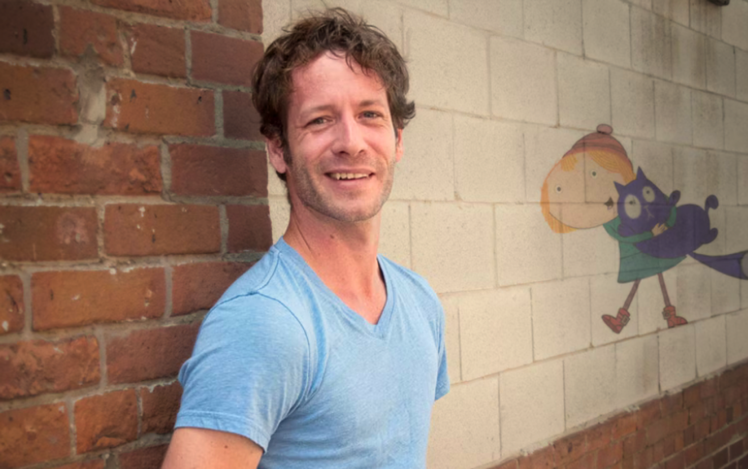 A photo of Cory Bobiak in front of a brick wall with an illustration of Peg + Cat on it