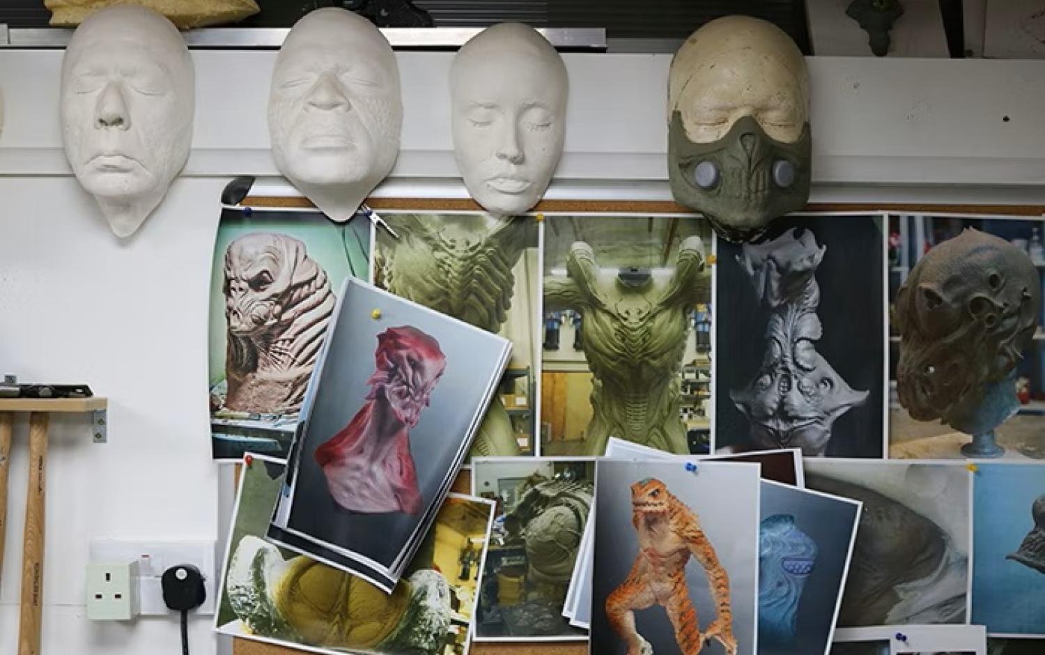 Wall display of creature design references and sculpted masks in a studio.