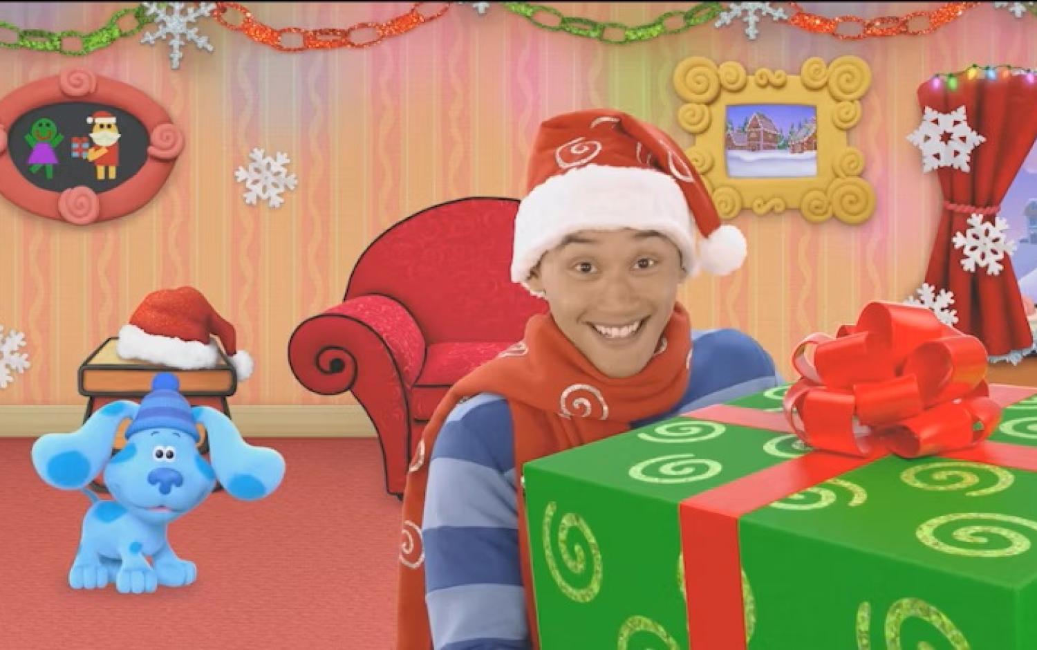 Blue and Josh wearing Santa hats in a festive room with holiday decorations and a large green gift box with a red bow.
