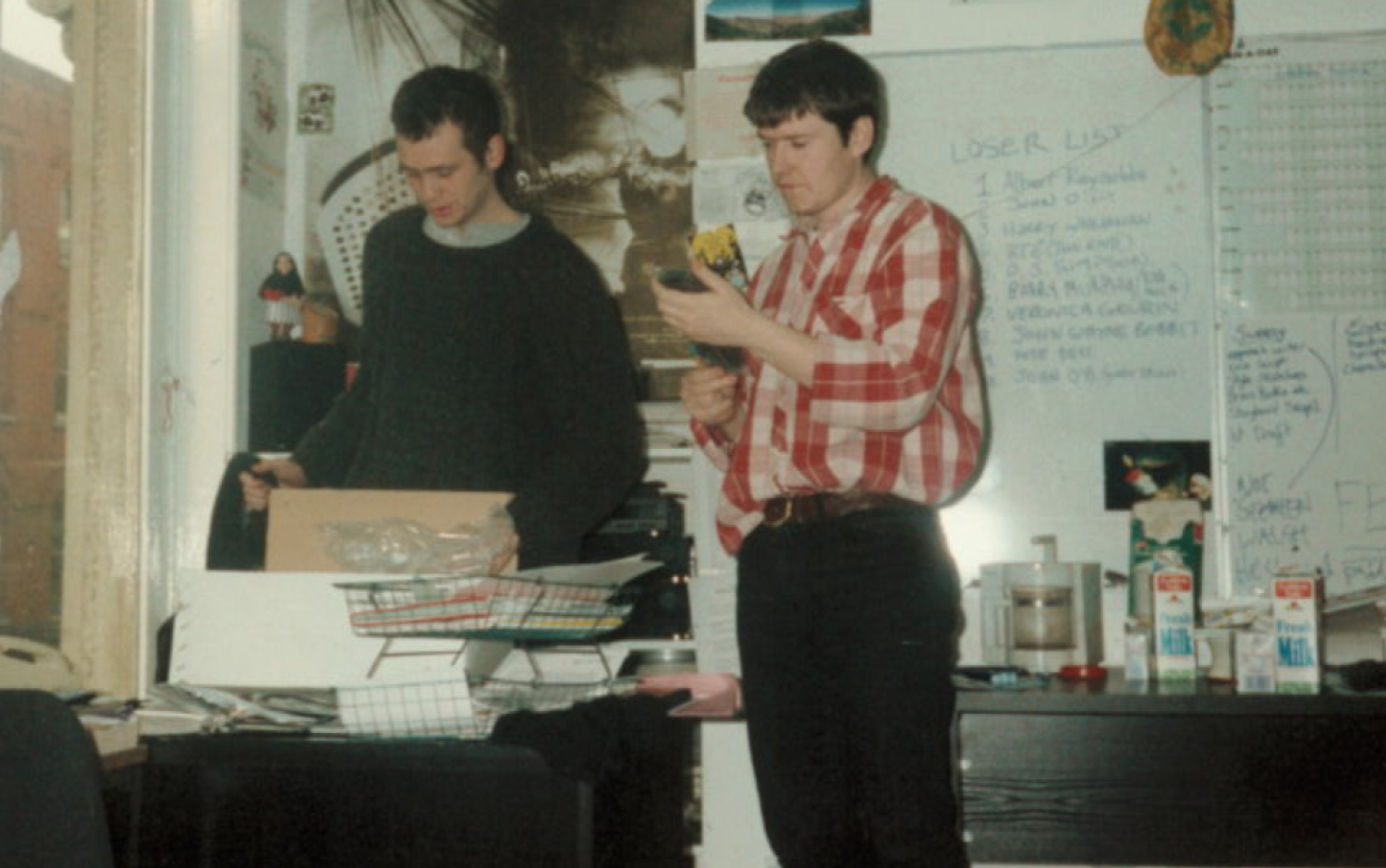 Two people standing in a cluttered office space with papers, boxes, and a whiteboard covered in notes.