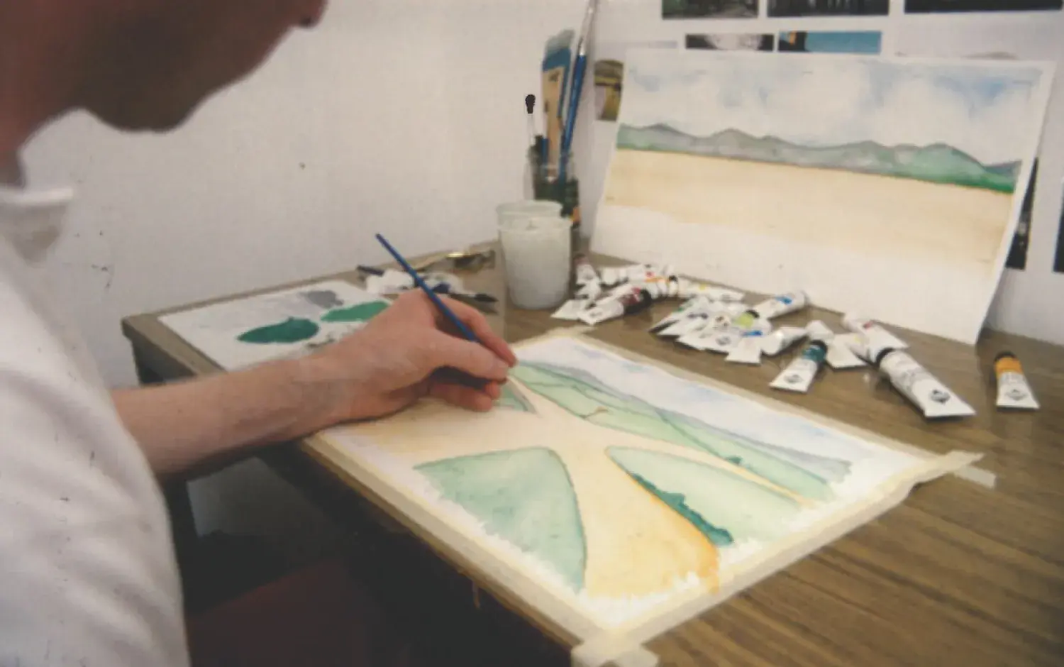 Close-up of an artist painting a watercolor landscape at a desk, surrounded by paint tubes and brushes.