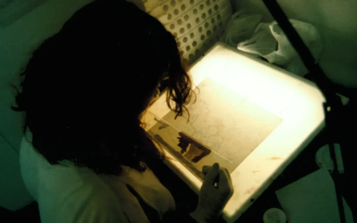 Person working at a lightbox, tracing or painting animation cels in a dimly lit studio.