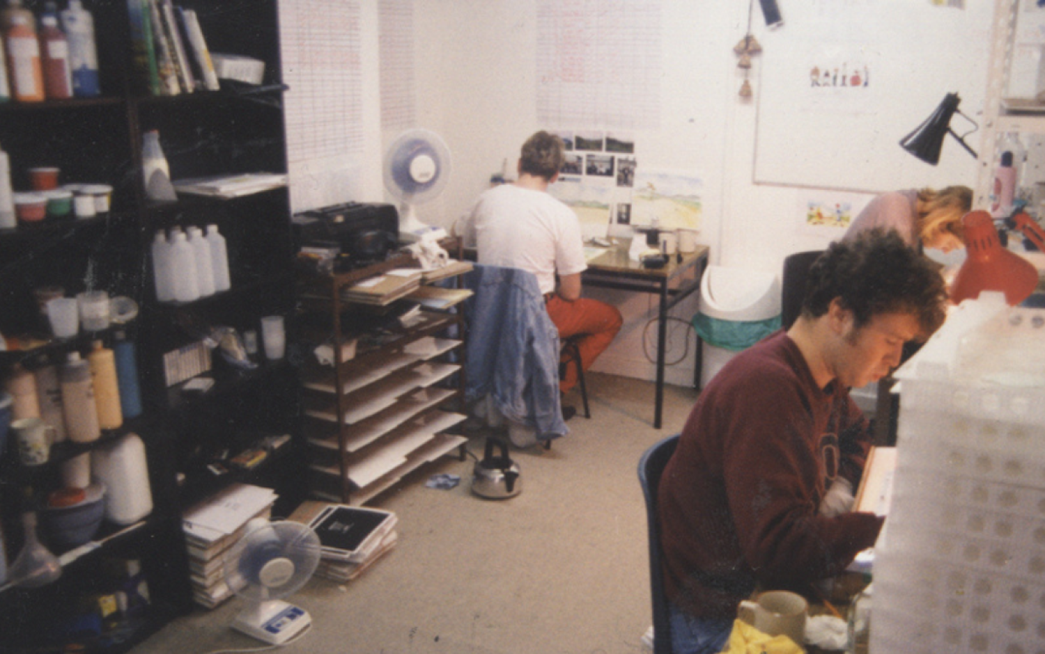 Animation studio workspace with shelves of supplies and three people seated at desks working on artwork.