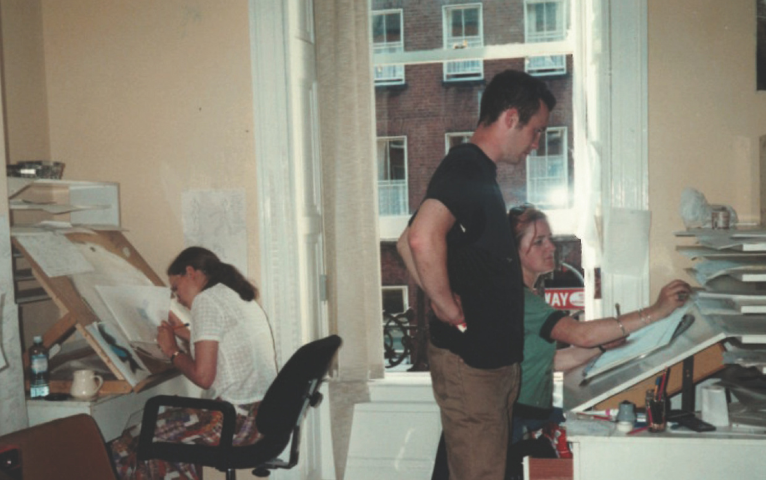 Studio scene with two animators at desks and another person standing, reviewing work near a large window.