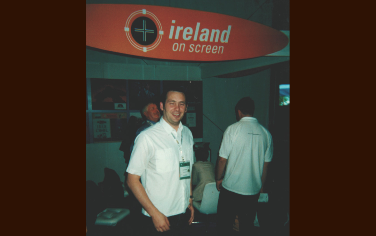 Exhibition booth with a red banner reading “Ireland on Screen”; people wearing white shirts with name badges stand beneath the sign.