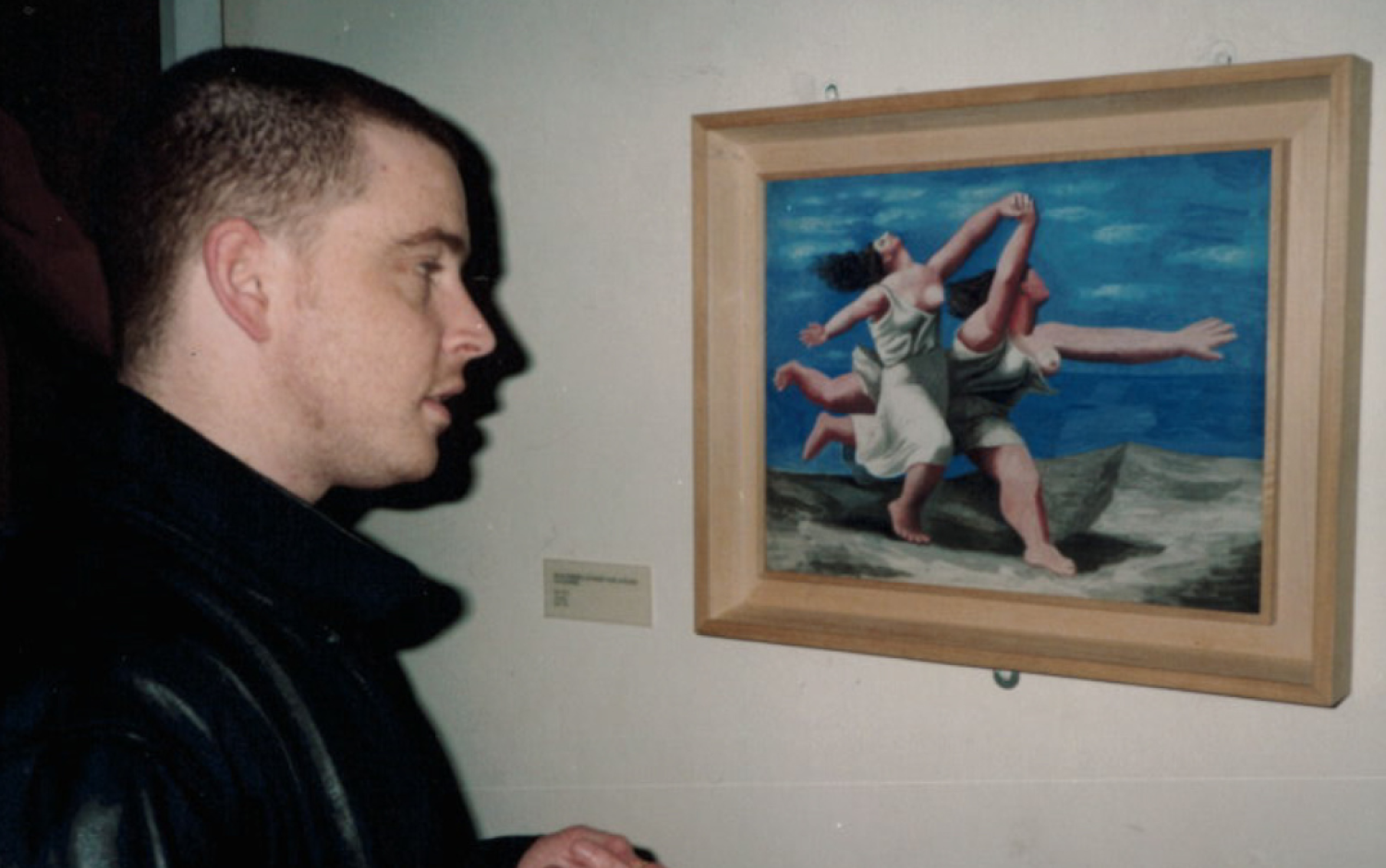 Person viewing a framed painting in an art gallery; the artwork depicts figures in dynamic poses against a blue sky.