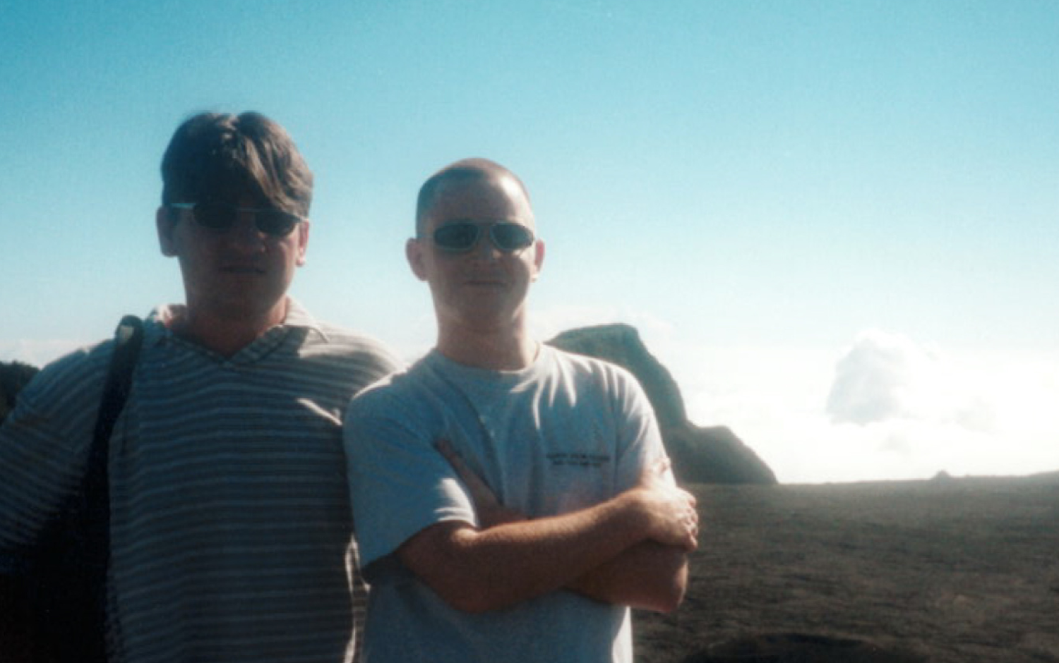 Two individuals standing outdoors on rocky terrain with a bright blue sky and clouds in the background.