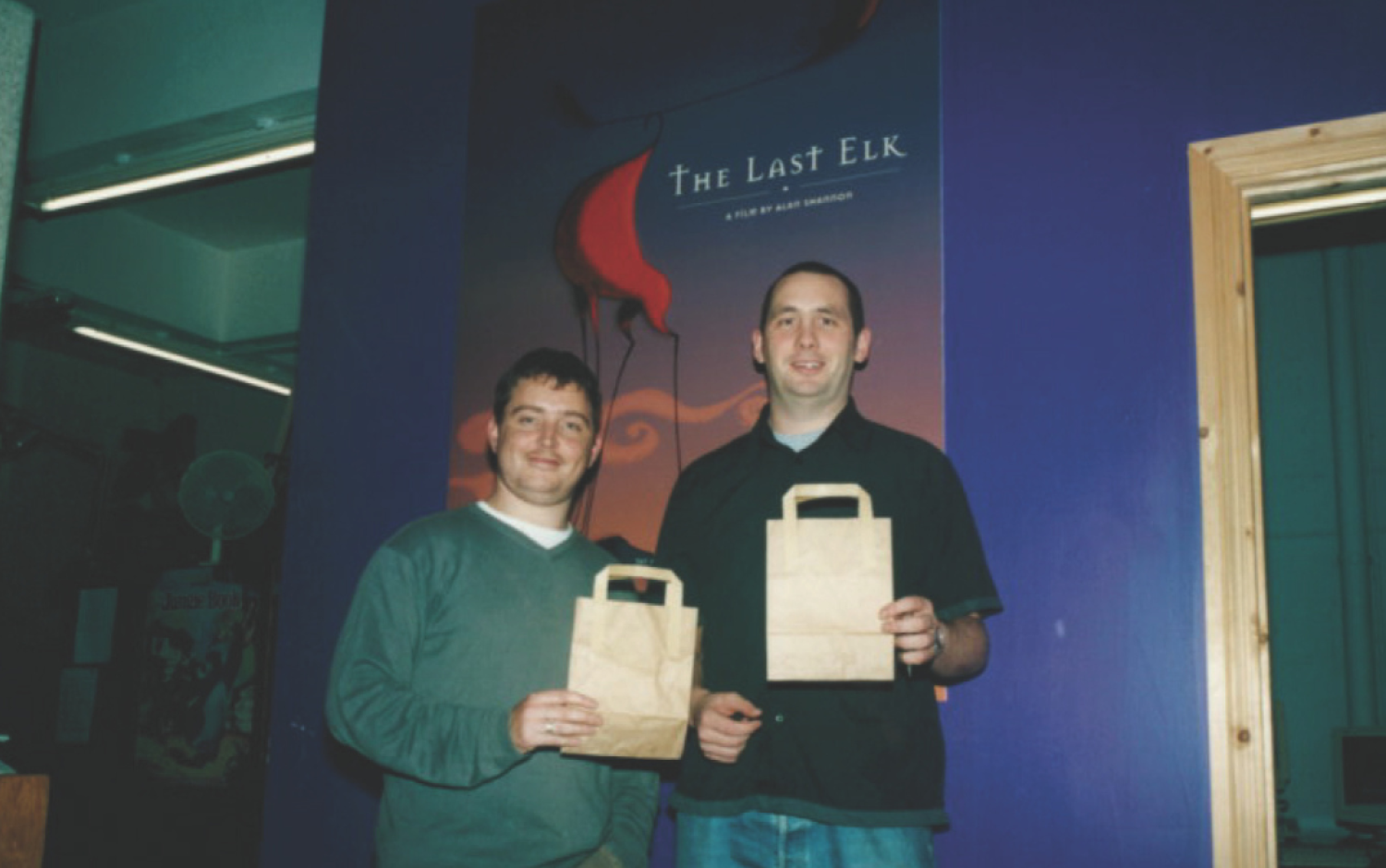 Two people standing in front of a purple wall with a poster for The Last Elk, each holding a small brown paper bag.