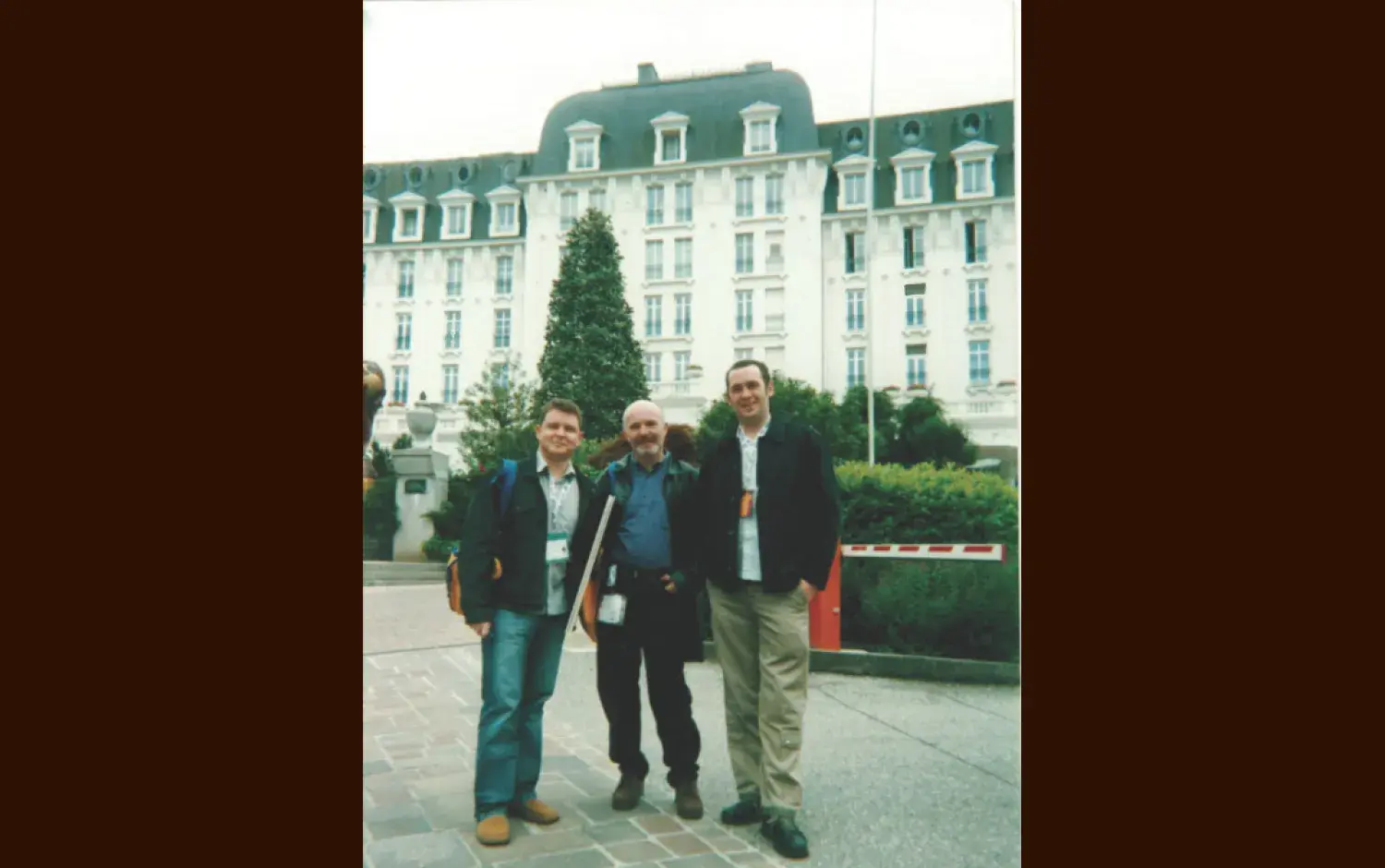 Three people standing outdoors in front of a large, ornate building with a mansard roof and landscaped grounds.