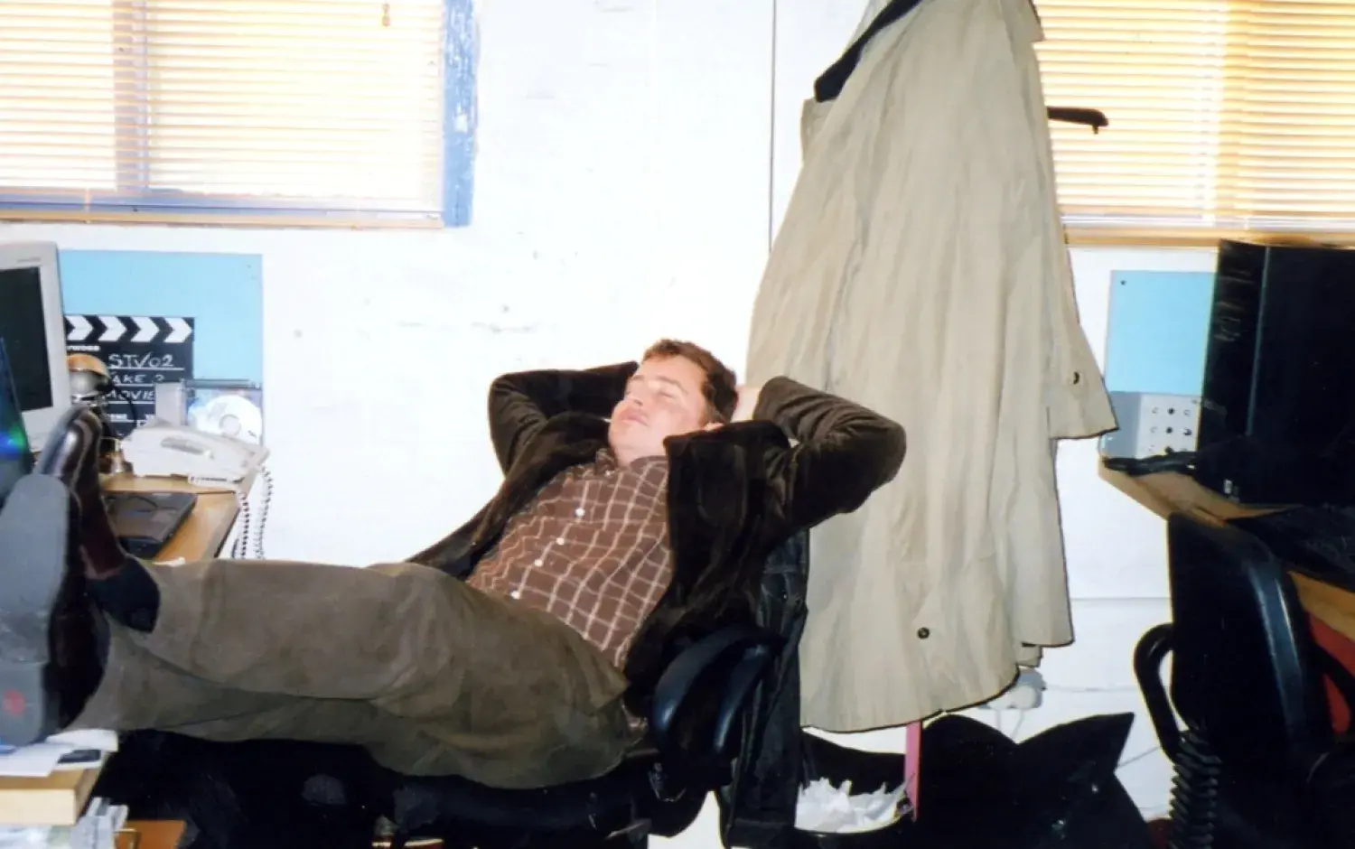 Person reclining in an office chair with feet propped on a desk, surrounded by computer equipment and a coat hanging nearby.