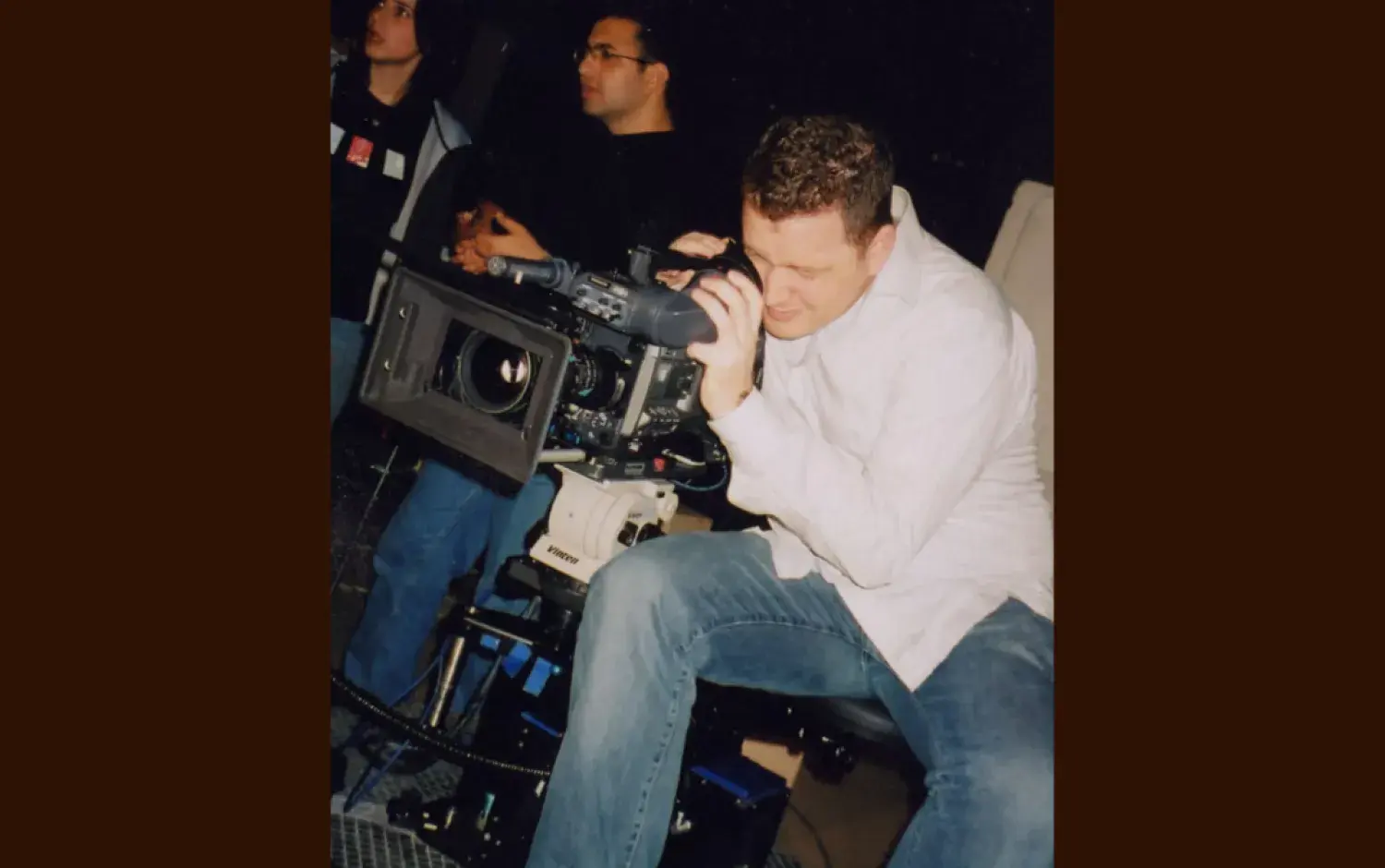Person operating a large professional video camera mounted on a tripod, with additional crew and equipment visible in the background.