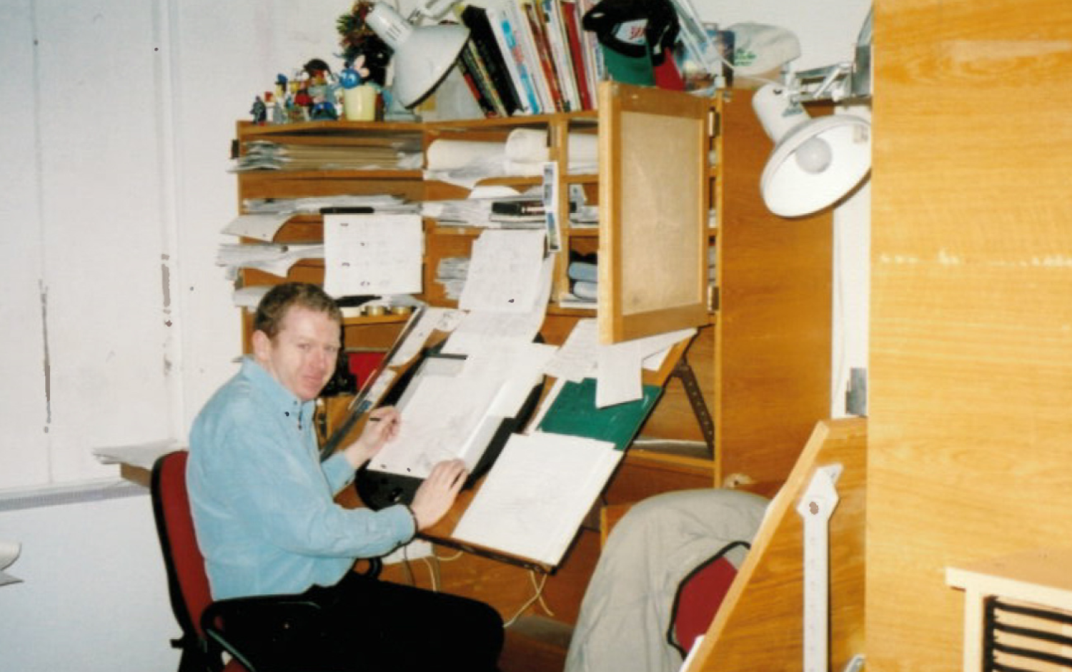 Animator seated at a wooden desk covered with papers and reference materials, working on hand-drawn animation sheets.