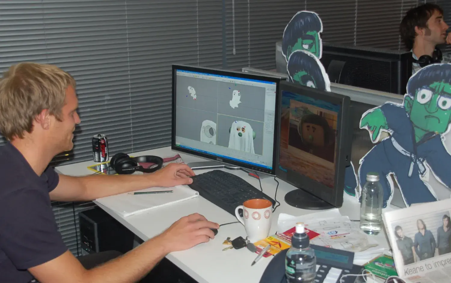 Person working at a dual-monitor setup with 3D character models on one screen and animation on the other; desk decorated with cut-out character art.