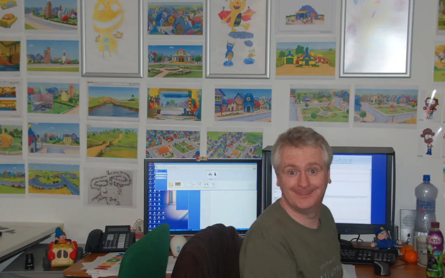 Person seated at a desk with two monitors, surrounded by colorful concept art pinned to the wall depicting animated characters and environments.