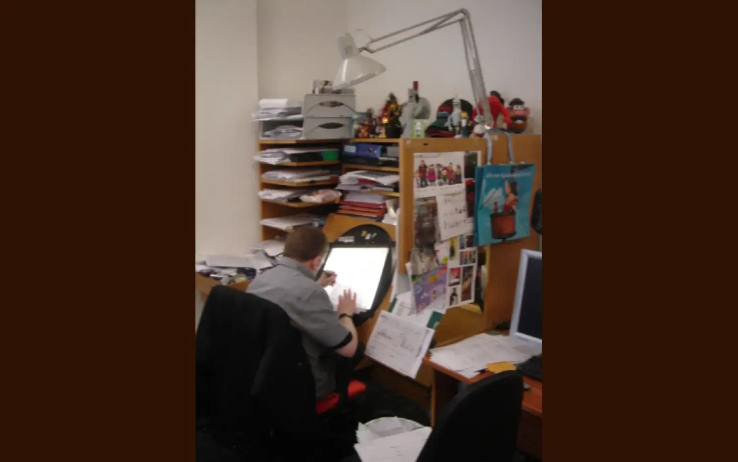 Animator seated at a cluttered desk using a lightbox to work on hand-drawn animation sheets; shelves behind are filled with papers and figurines.