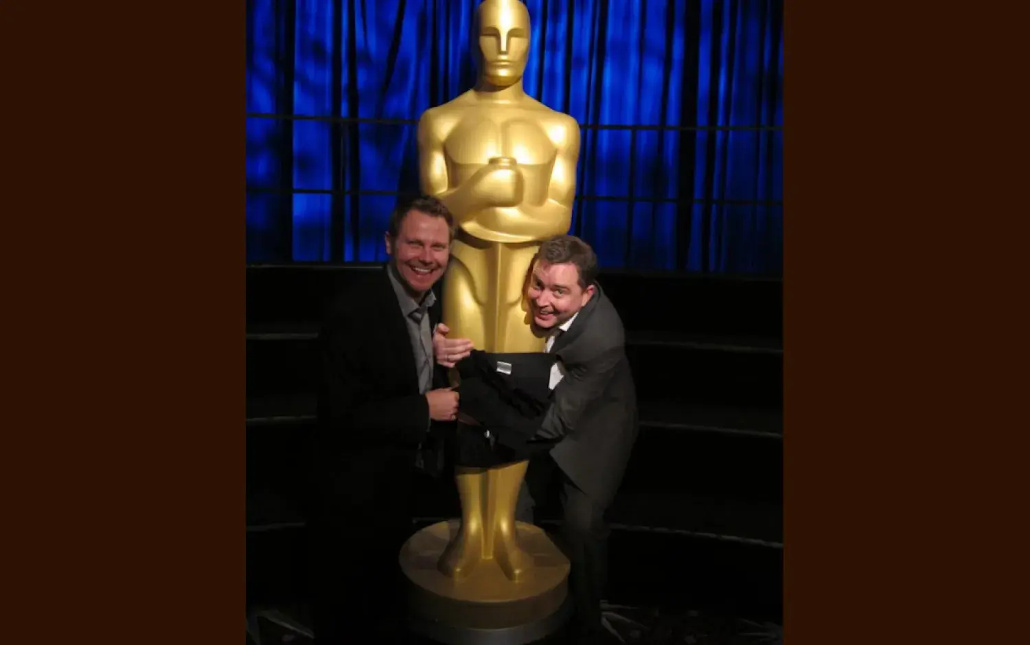 Two people in formal suits playfully posing with a large golden Oscar statue on a stage.