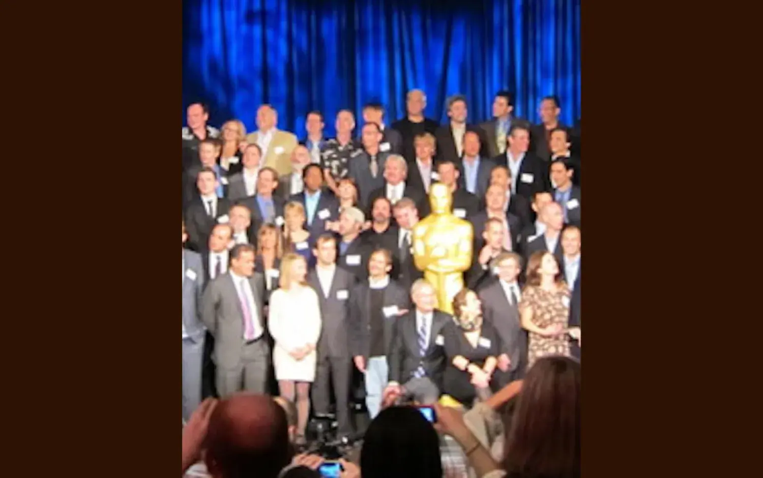 Group photo of Academy Awards nominees on stage with a large golden Oscar statue in front of a blue curtain.