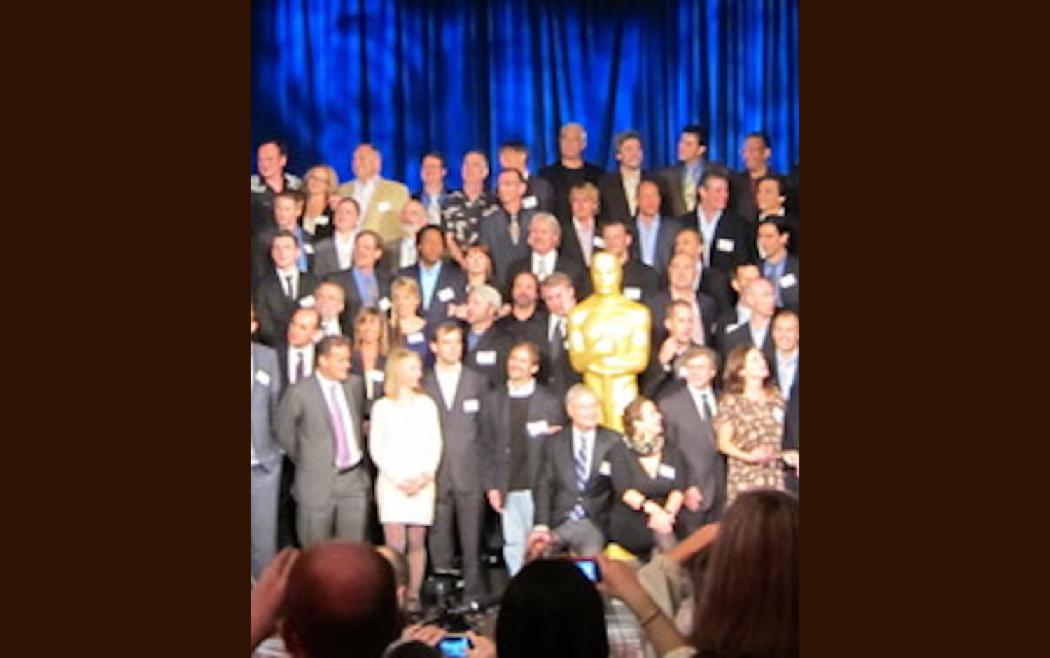 Group photo of Academy Awards nominees on stage with a large golden Oscar statue in front of a blue curtain.